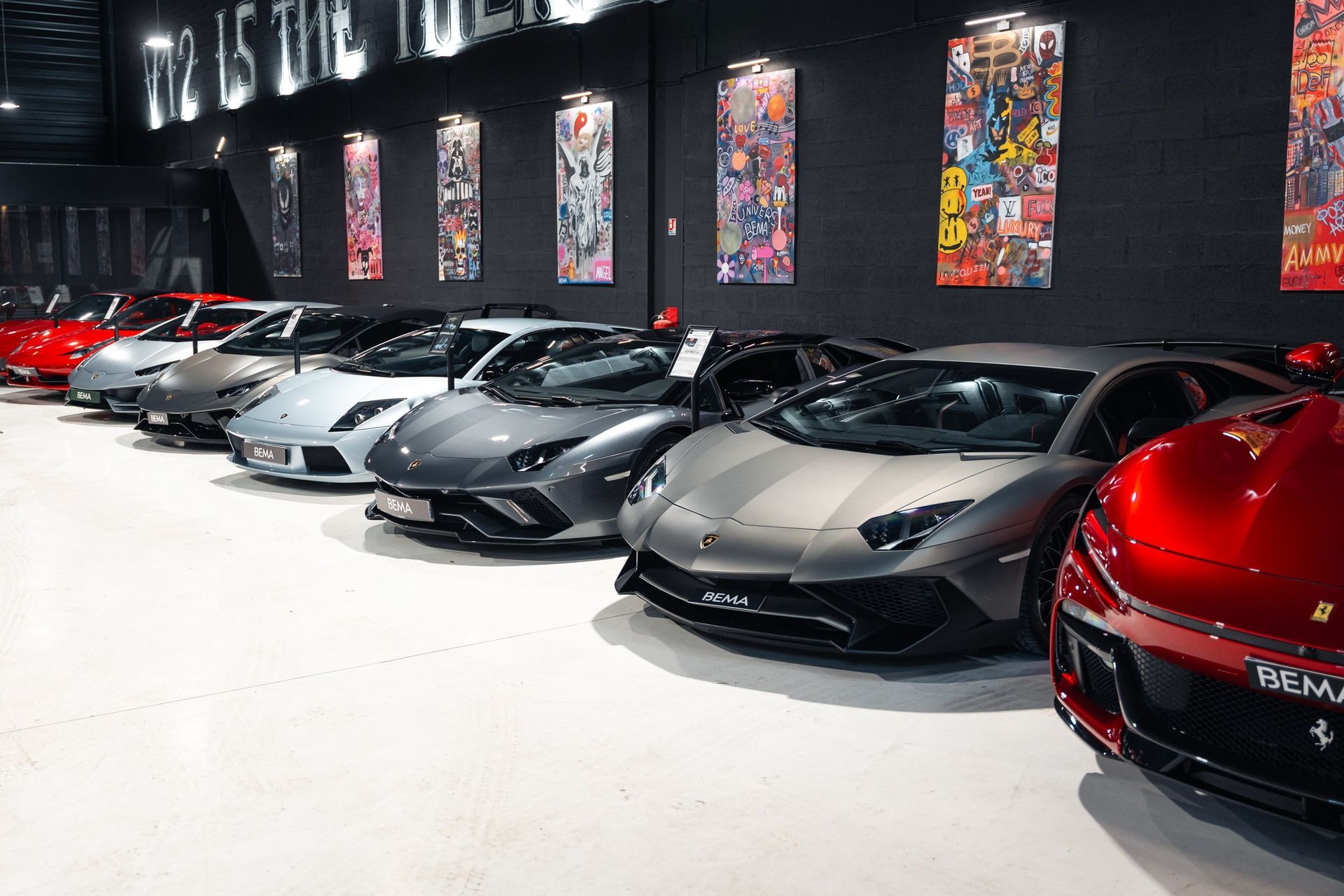 Des voitures de luxe, dont des Lamborghini et une Ferrari, alignées dans une salle d'exposition avec des œuvres d'art sur les murs.
