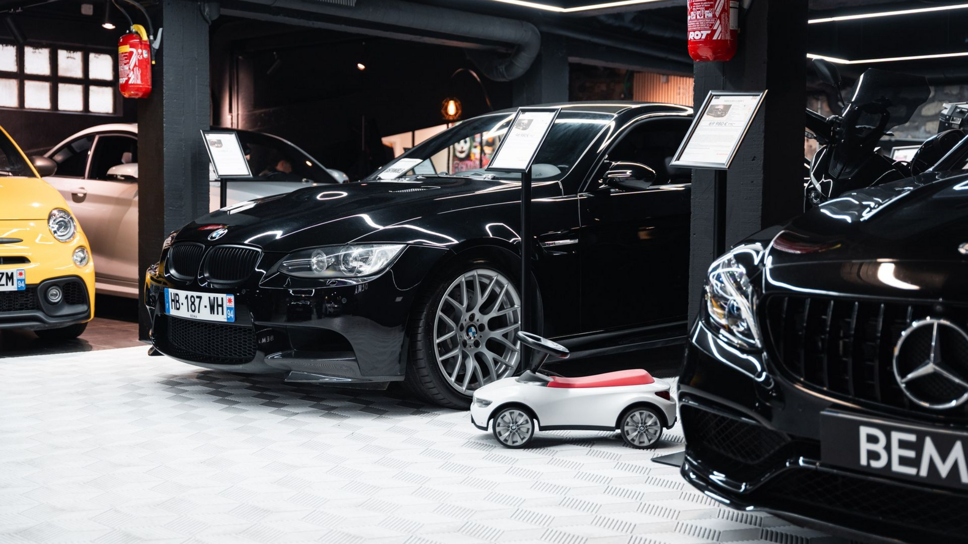 Voitures dans un showroom : BMW noire, Mercedes et Fiat jaune. Une petite voiture d'enfant est devant.