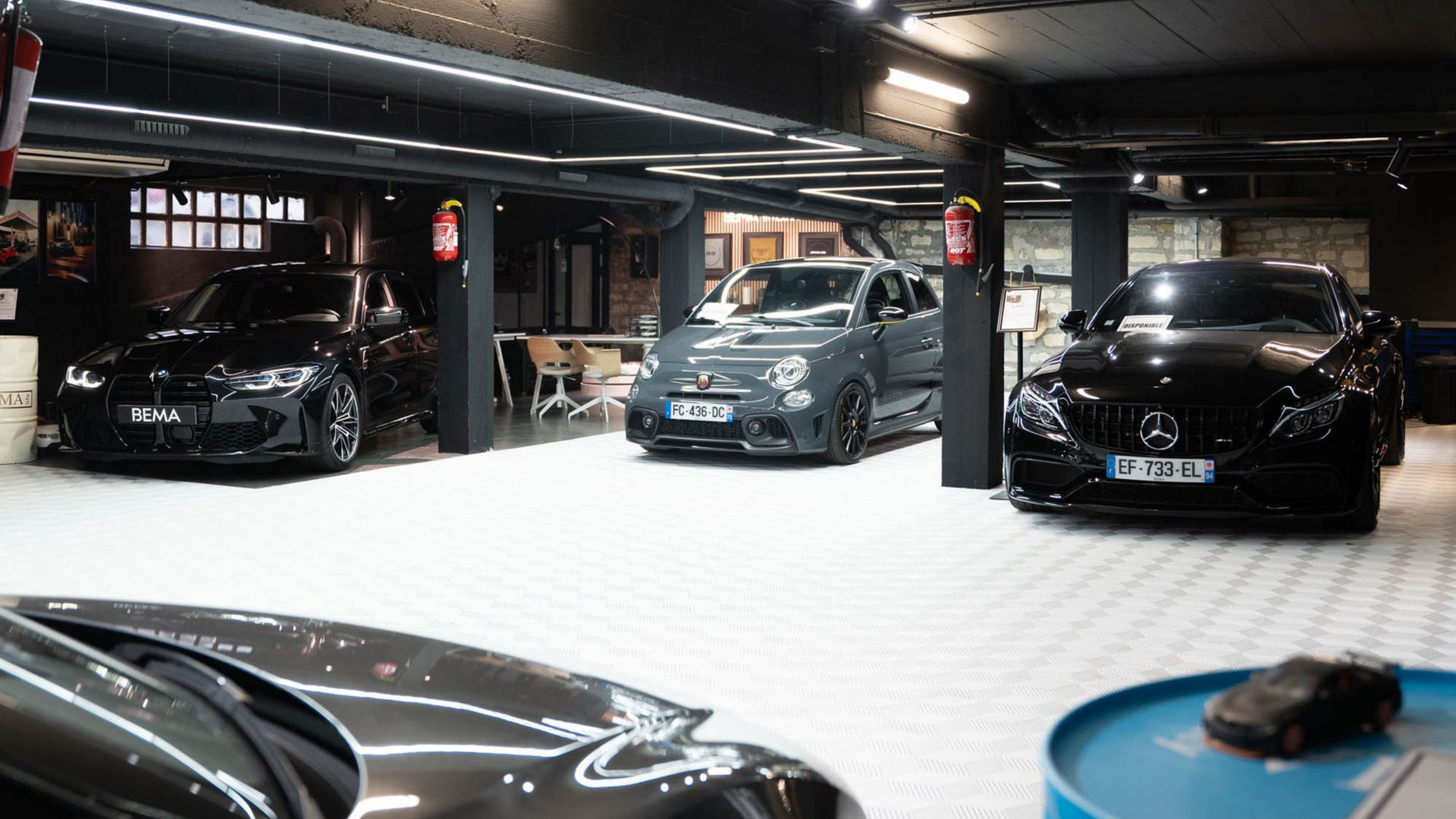 Garage sombre avec plusieurs voitures, dont une voiture de sport noire au premier plan, sous des lumières vives au plafond.