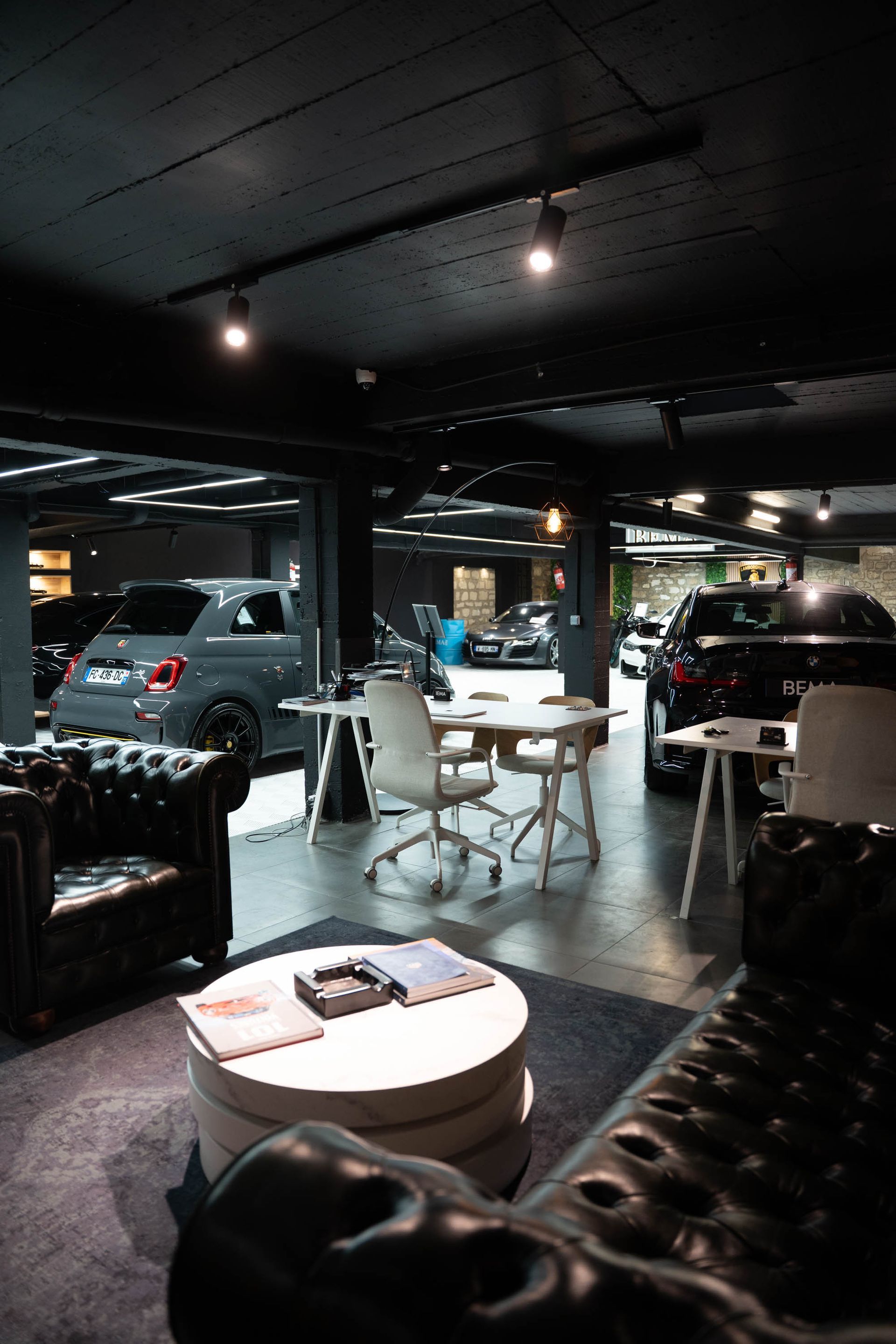 Intérieur de salle d'exposition sombre avec des voitures, des bureaux blancs et un canapé en cuir.