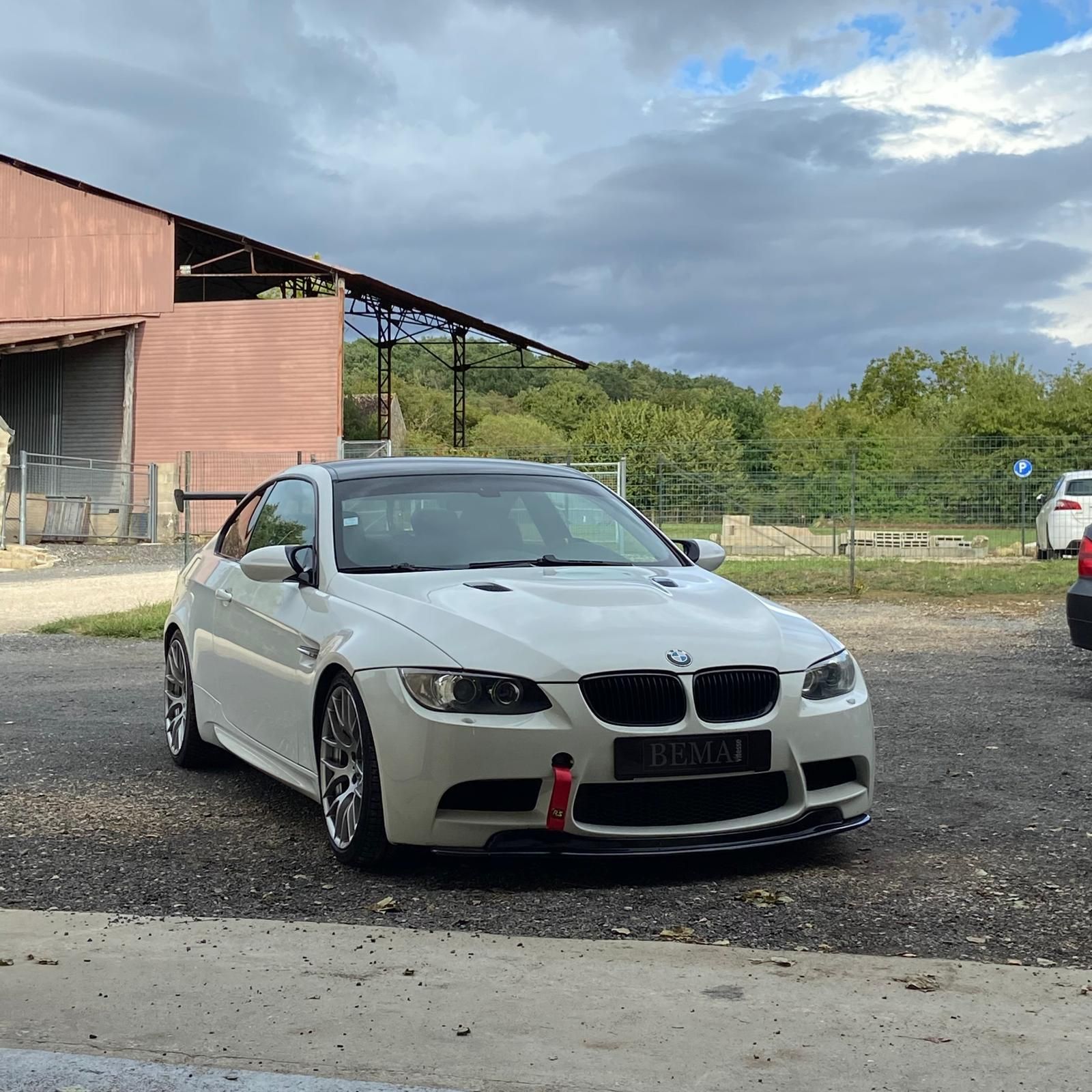 Coupé BMW M3 blanc garé à l'extérieur d'un bâtiment rustique sous un ciel nuageux.