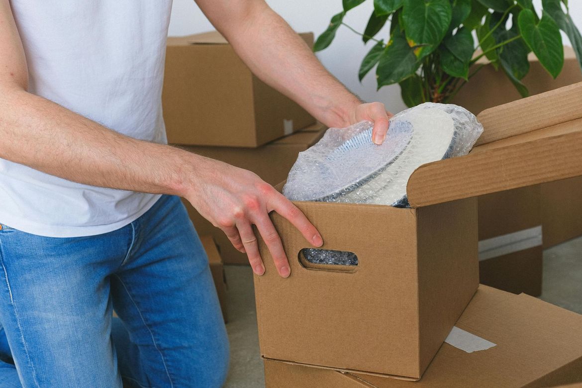Persona empacando zapatos en una caja de cartón, rodeada de más cajas, probablemente mudándose.