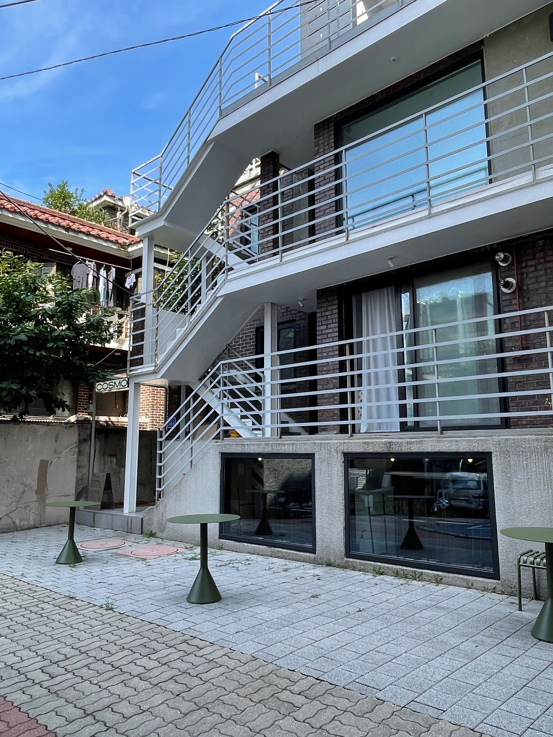 PIPE Hostel. Modern white building with external stairs, brick facade, and small outdoor tables.