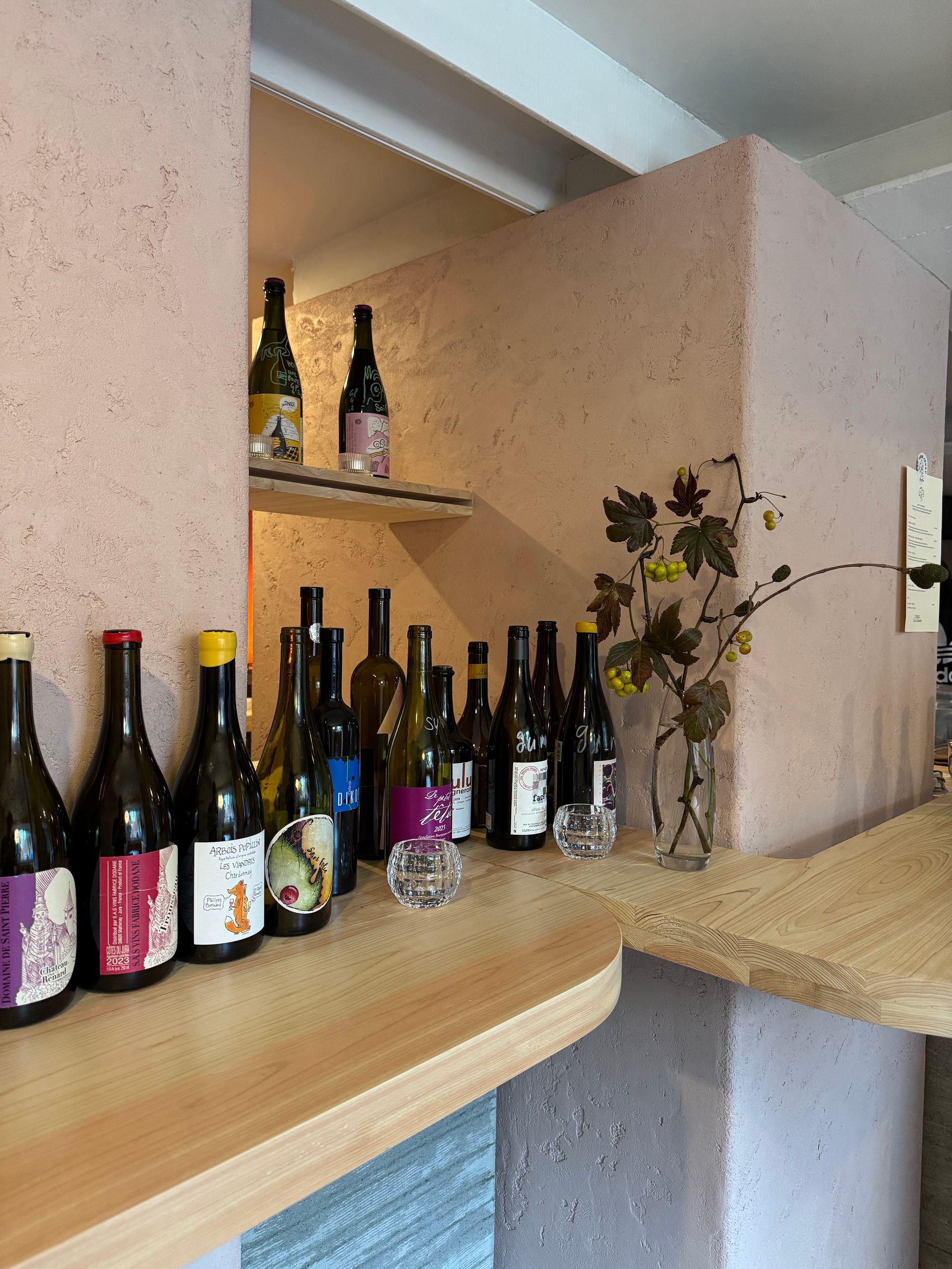 Bottles of wine on a counter, displayed against a pink wall with a shelf holding more bottles. COSMO Wine Bar