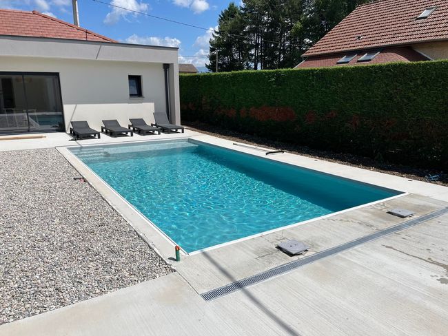 Une piscine extérieure à l'eau bleue, entourée de gravier, d'une terrasse en béton et d'une haute haie, jouxte une maison blanche.
