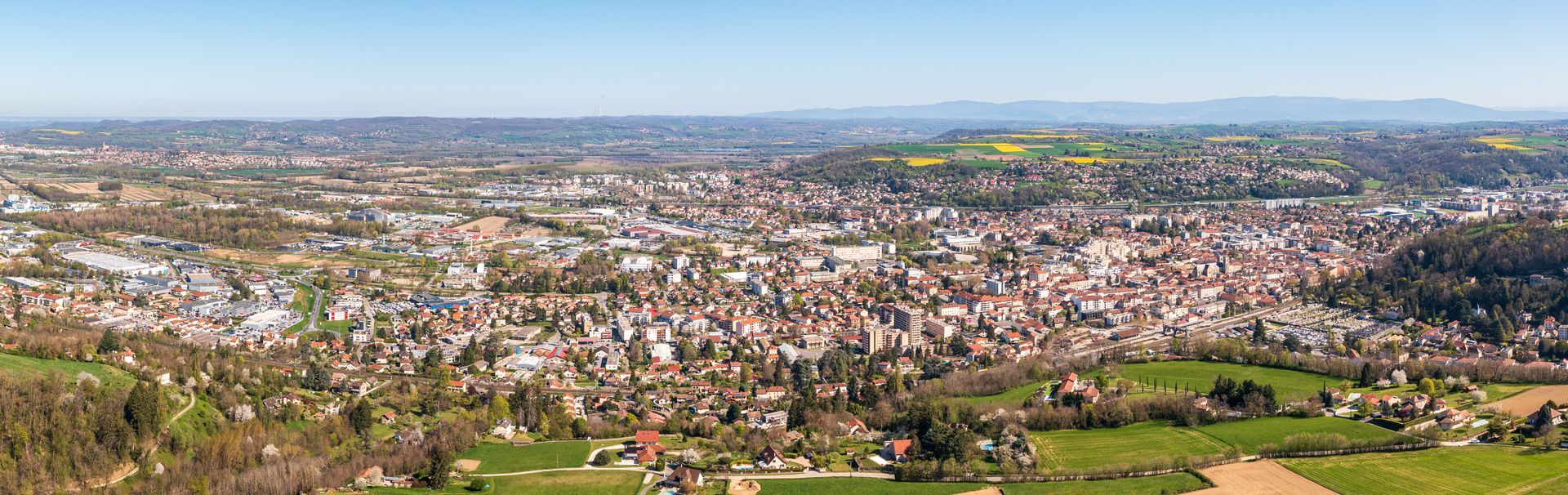 Vue panoramique en plongée d'une ville dense nichée dans une vallée aux collines verdoyantes et sous un ciel d'un bleu éclatant.
