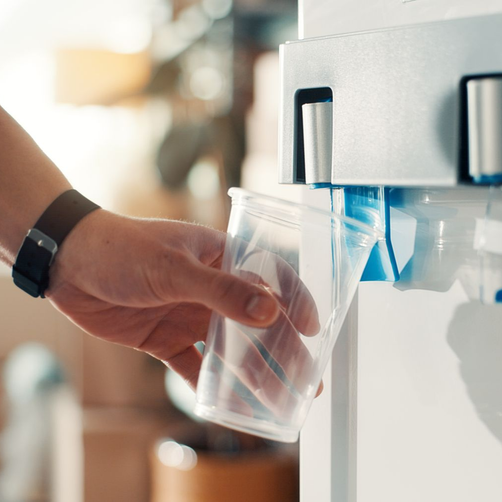 Une main tient un gobelet en plastique transparent sous un distributeur d'eau pour le remplir.