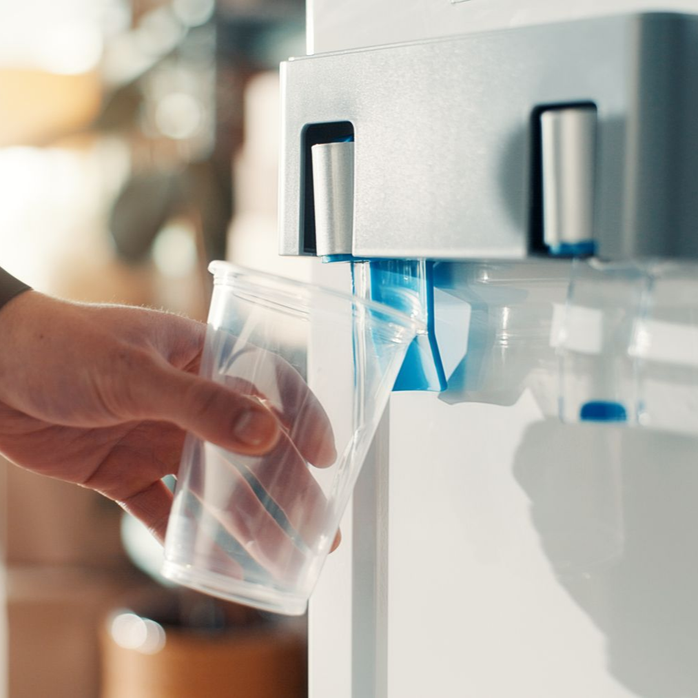 Une main tient un gobelet en plastique sous un distributeur d'eau fixé sur une surface blanche, recueillant l'eau provenant du bec bleu.