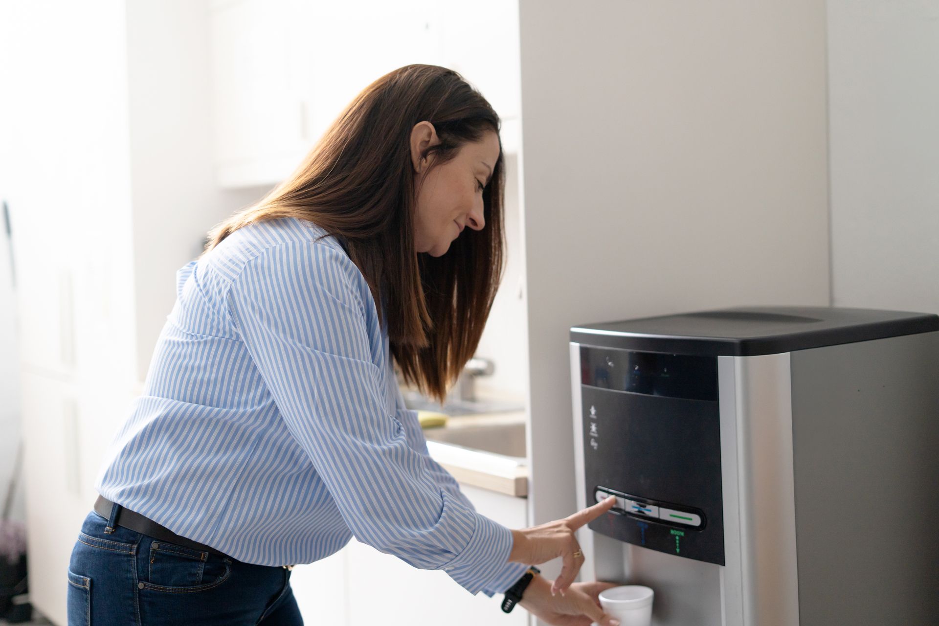 Dans une cuisine moderne et lumineuse, une personne vêtue d'une chemise à rayures bleu clair remplit un gobelet blanc à une fontaine à eau.