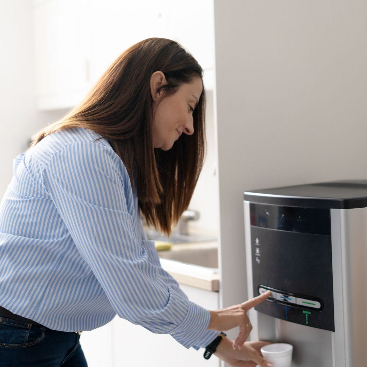 Dans une cuisine, une personne vêtue d'une chemise rayée à boutons appuie sur un bouton d'un distributeur d'eau tout en tenant une tasse.