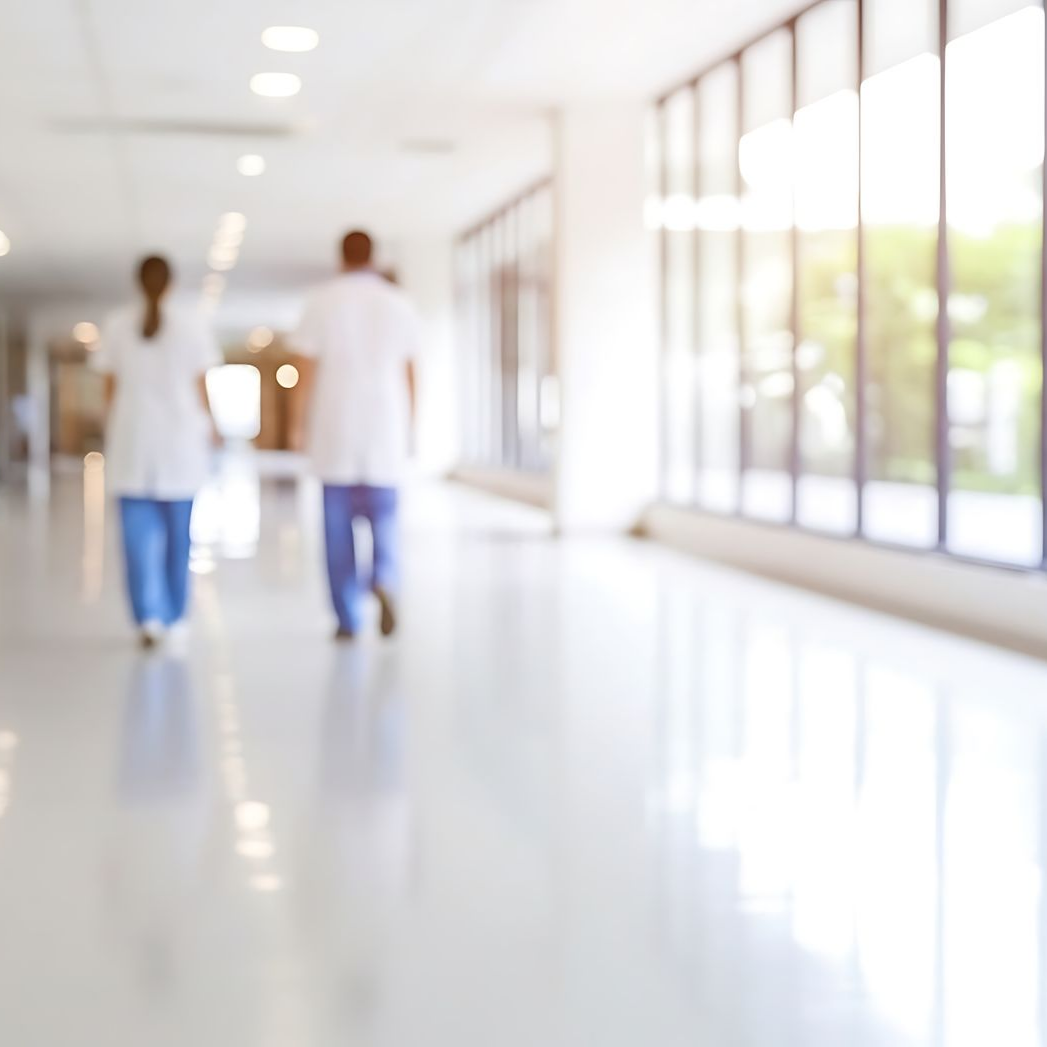Deux professionnels de santé en blouse médicale descendent un couloir d'hôpital lumineux et flou, doté de grandes fenêtres.