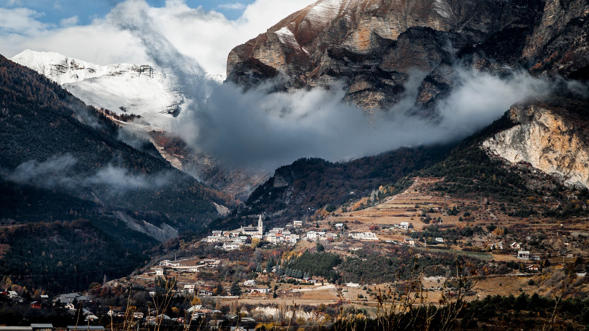 Un petit village de montagne niché dans une vallée luxuriante et brumeuse, au pied d'imposants pics rocheux enneigés.