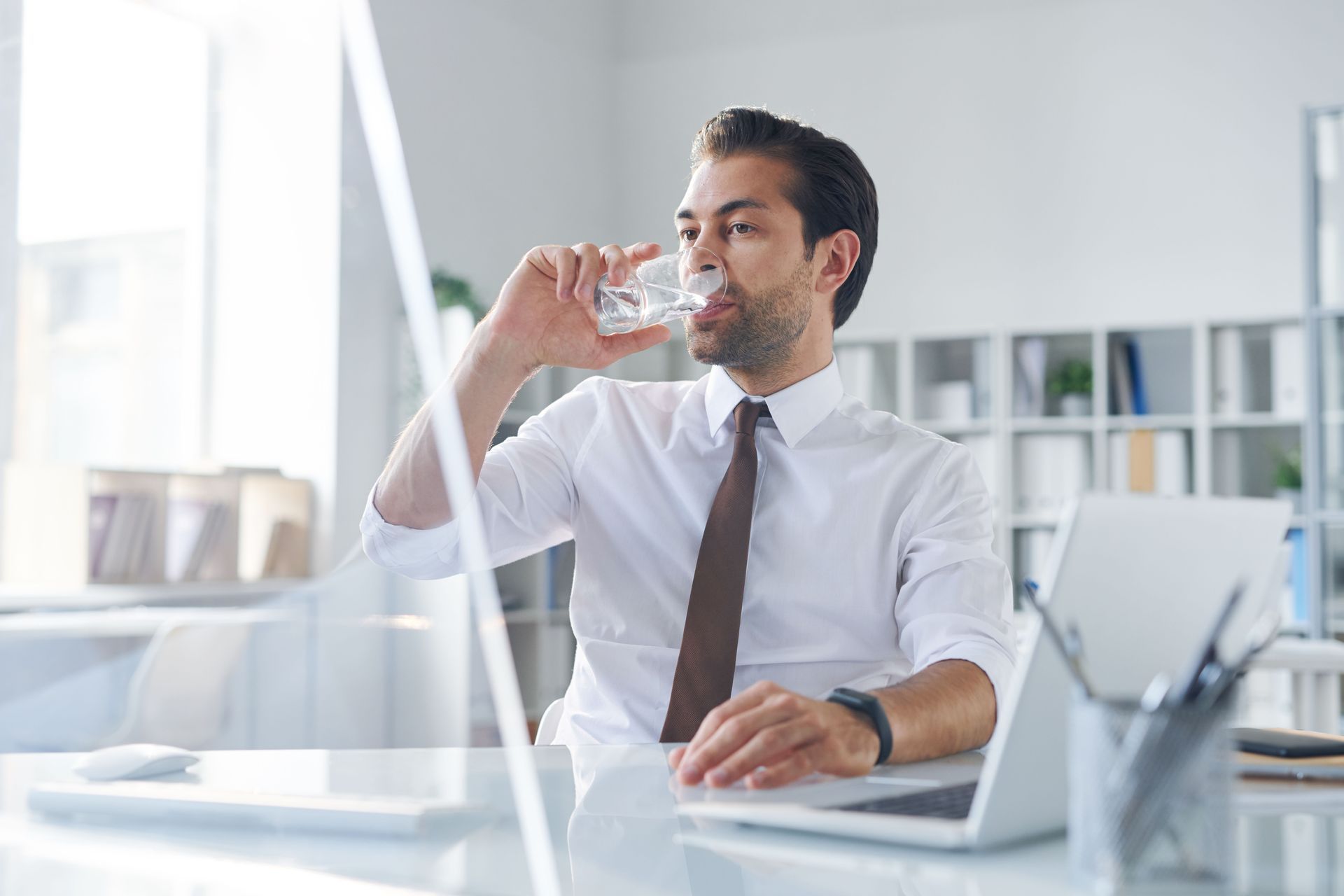 Une personne en chemise blanche et cravate boit un verre d'eau assise à un bureau dans un bureau moderne et lumineux.