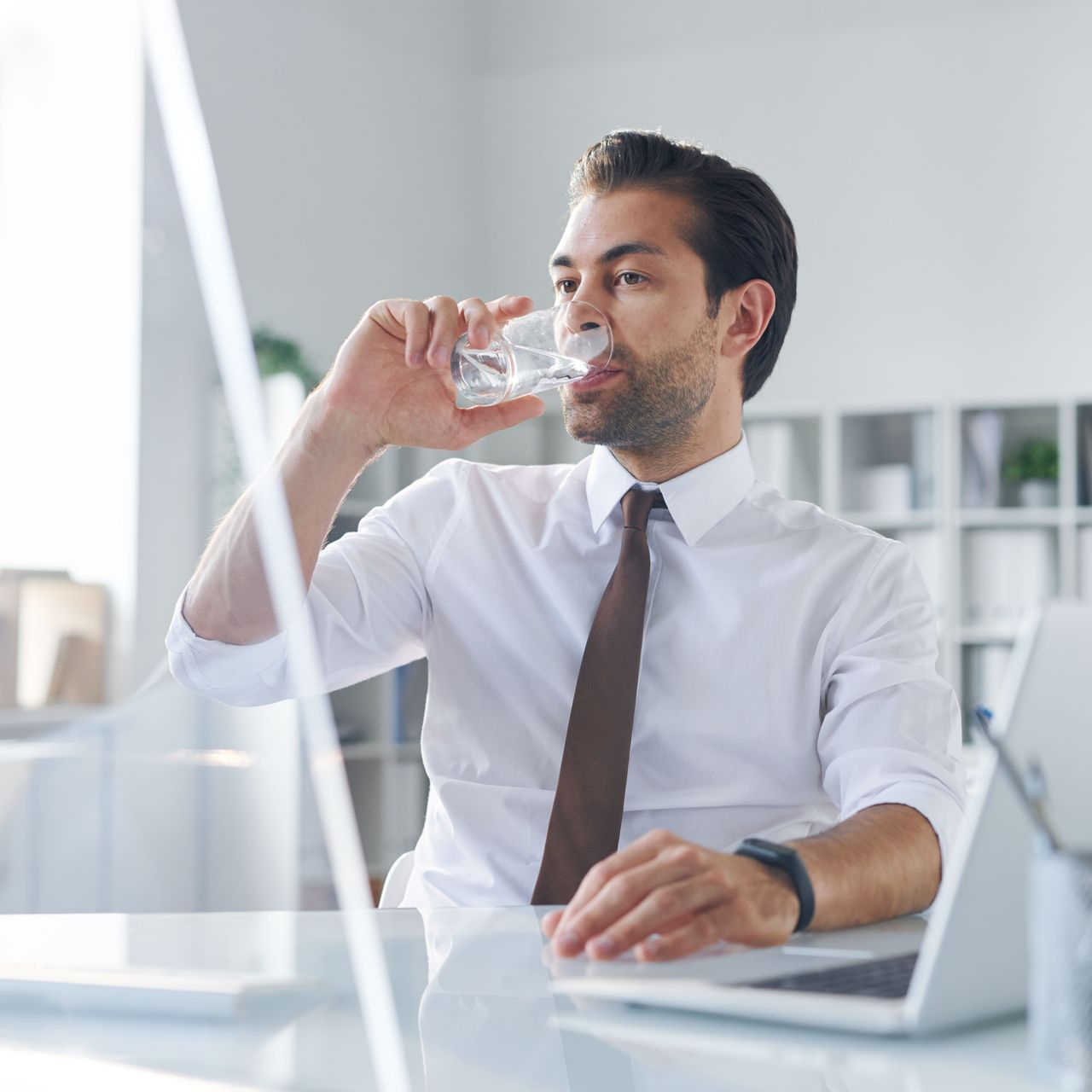 Dans un bureau lumineux, une personne vêtue d'une chemise blanche et d'une cravate marron est assise à un bureau et boit de l'eau dans un verre.