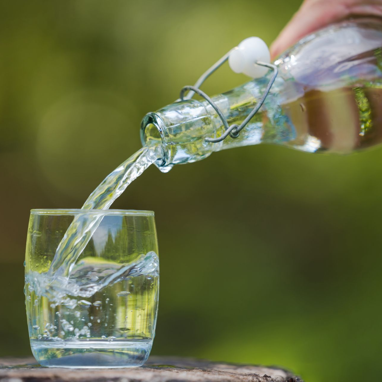 Une main verse de l'eau d'une bouteille en verre à bouchon mécanique dans un verre, sur un fond vert flou.