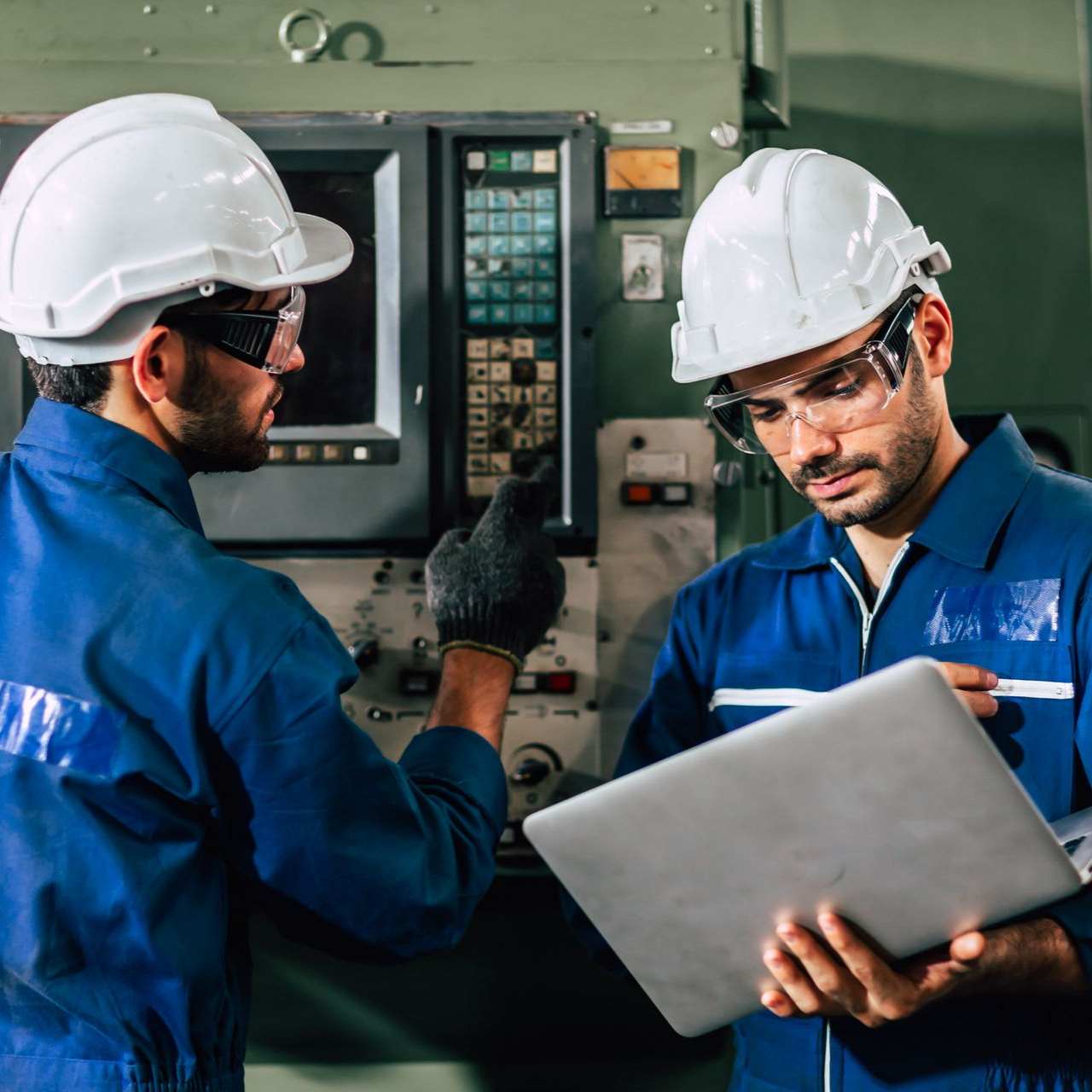 Deux techniciens en uniforme bleu, casques de chantier et lunettes de sécurité utilisent un ordinateur portable et les commandes d'une machine dans une usine.