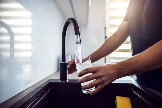 Une personne remplit un verre transparent d'eau provenant d'un robinet de cuisine moderne à la finition sombre, placé au-dessus d'un évier sombre.