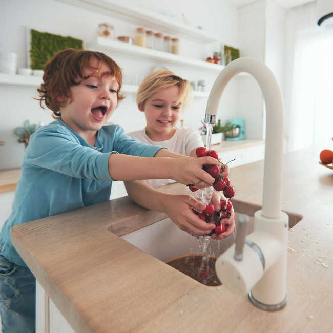 Dans une cuisine moderne et lumineuse, deux enfants lavent joyeusement une grappe de cerises rouges sous le robinet.