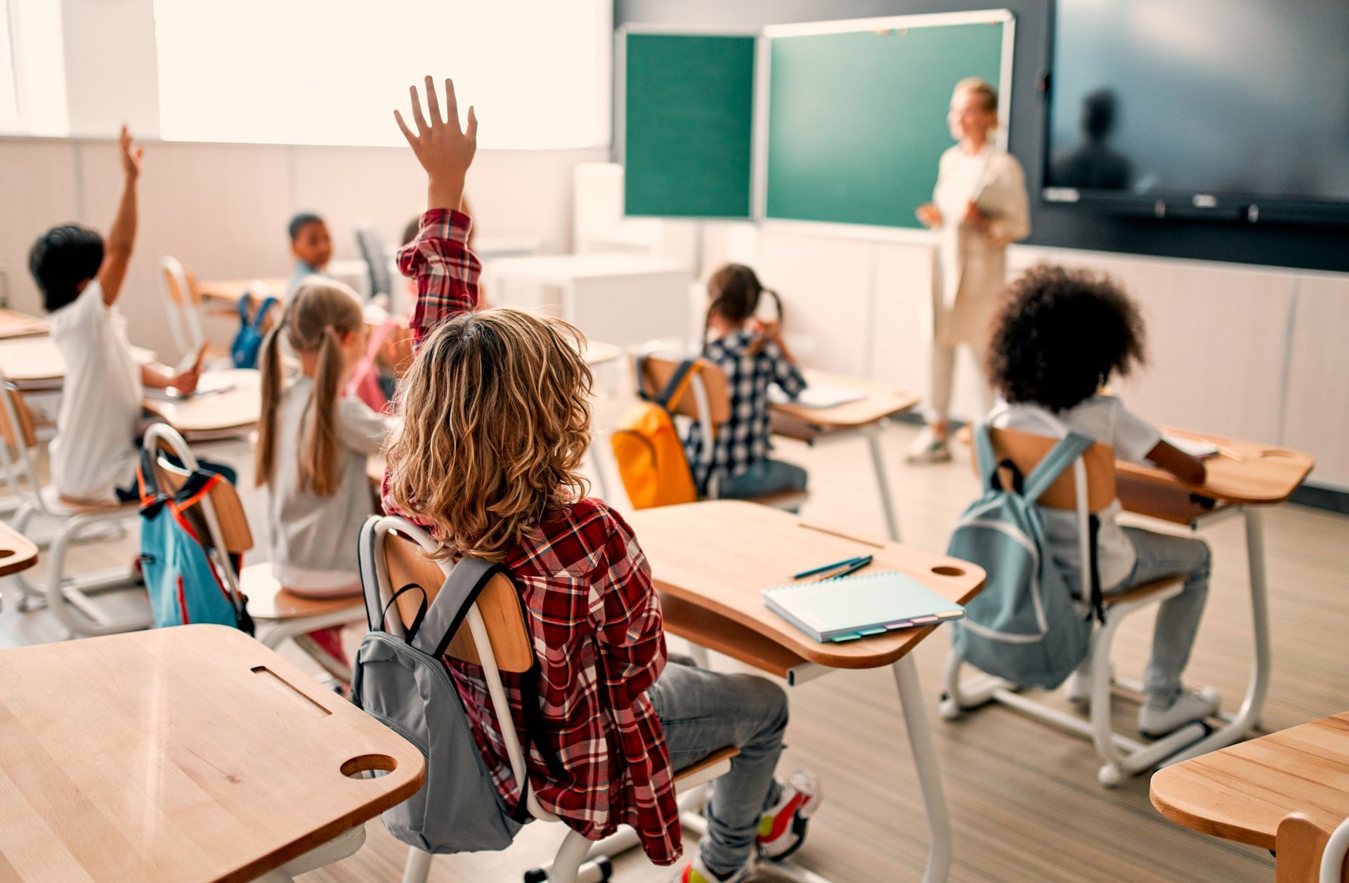 Des élèves dans une salle de classe, l'un d'eux levant la main vers un professeur debout près d'un tableau noir.
