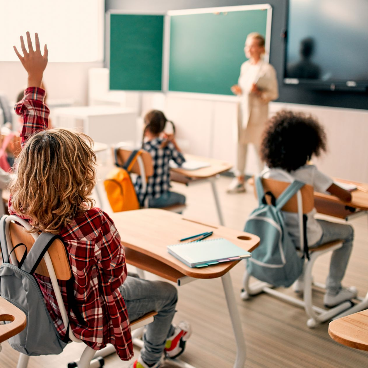 Des élèves dans une salle de classe, un enfant levant la main tandis qu'une enseignante se tient près d'un tableau blanc en arrière-plan.