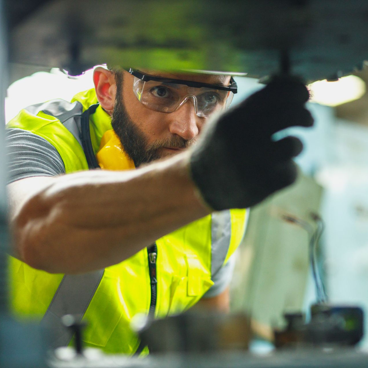 Un ouvrier concentré, vêtu d'un gilet jaune haute visibilité et de lunettes de sécurité, répare soigneusement des machines dans un atelier.