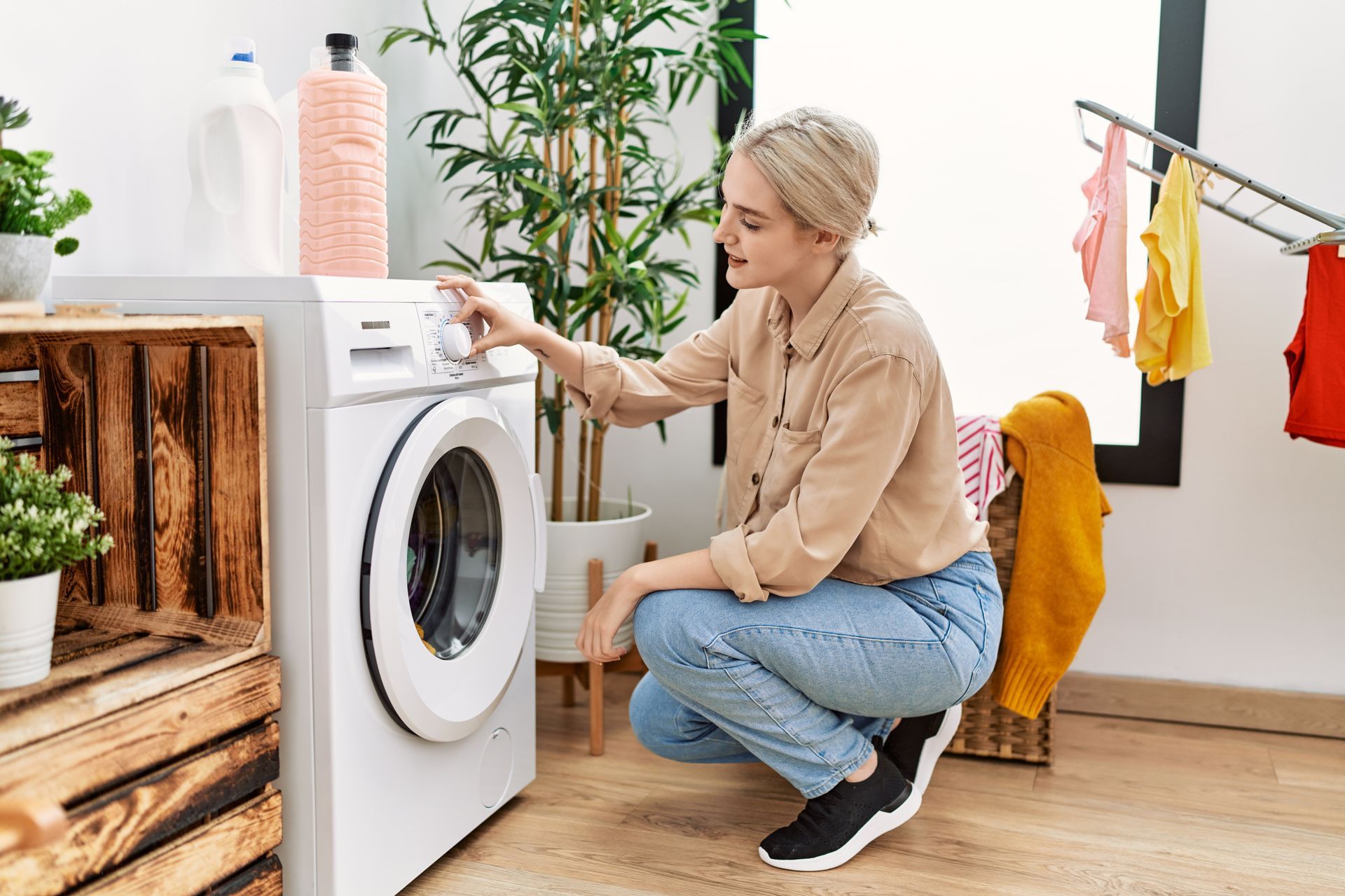 Une personne en chemise beige et jean est accroupie pour régler les paramètres d'une machine à laver blanche dans une buanderie lumineuse.