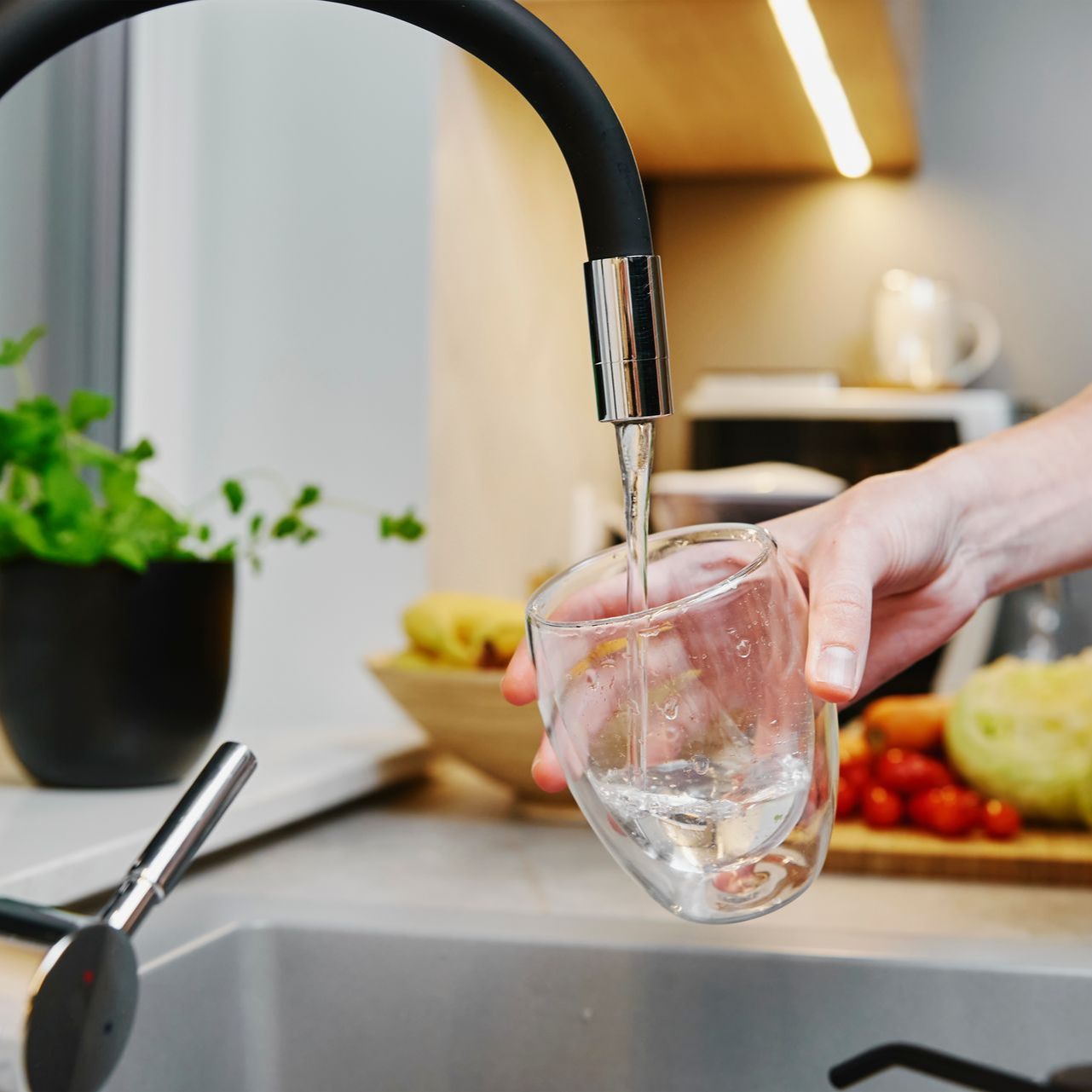 Dans une cuisine moderne et lumineuse, avec une plante et des aliments en arrière-plan, un robinet verse de l'eau dans un verre tenu par une main.