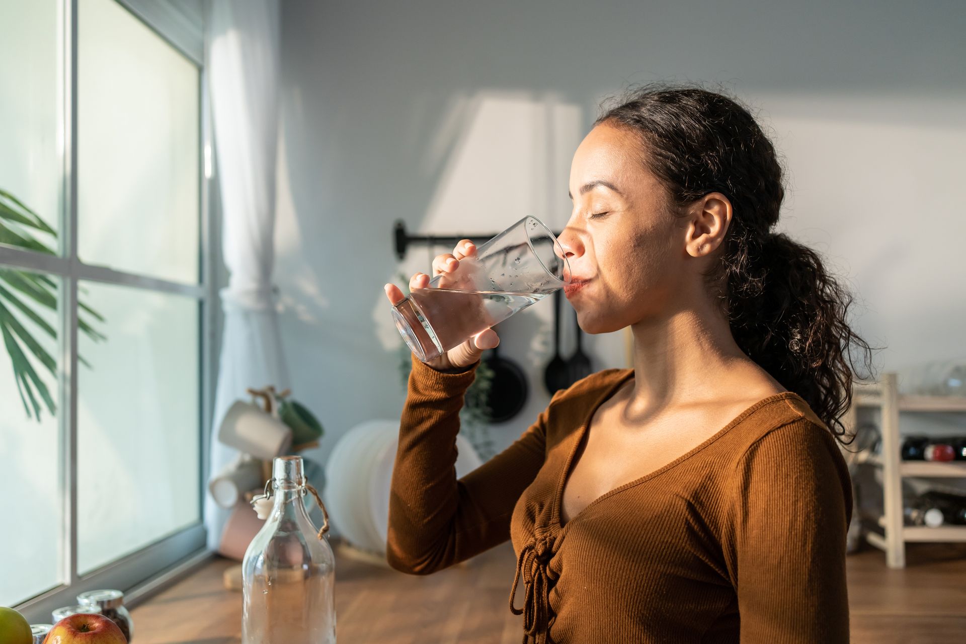 Une personne vêtue d'une chemise brune boit dans un verre d'eau près d'une fenêtre, son visage éclairé par la chaude lumière du soleil.