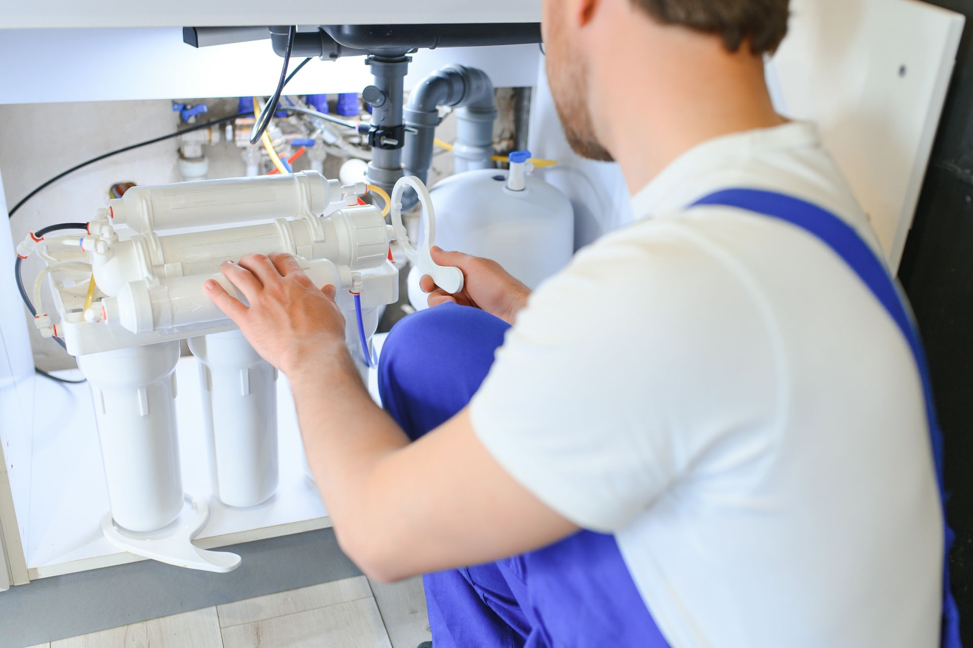 Un technicien en chemise blanche et salopette bleue installe un système de filtration d'eau sous l'évier.