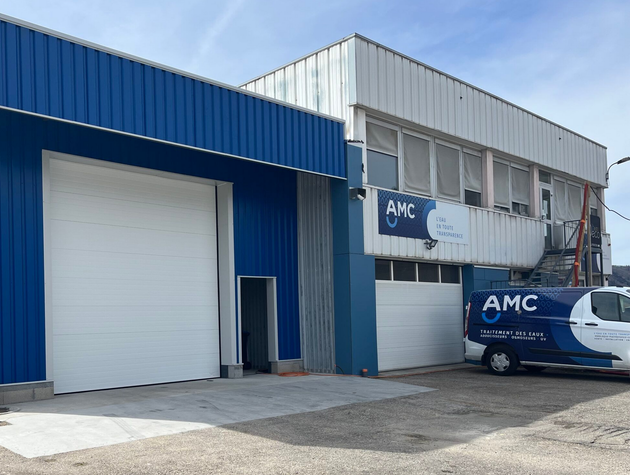 Un entrepôt industriel bleu et blanc avec une grande porte sectionnelle blanche et une camionnette de travail aux couleurs de la marque garée devant.