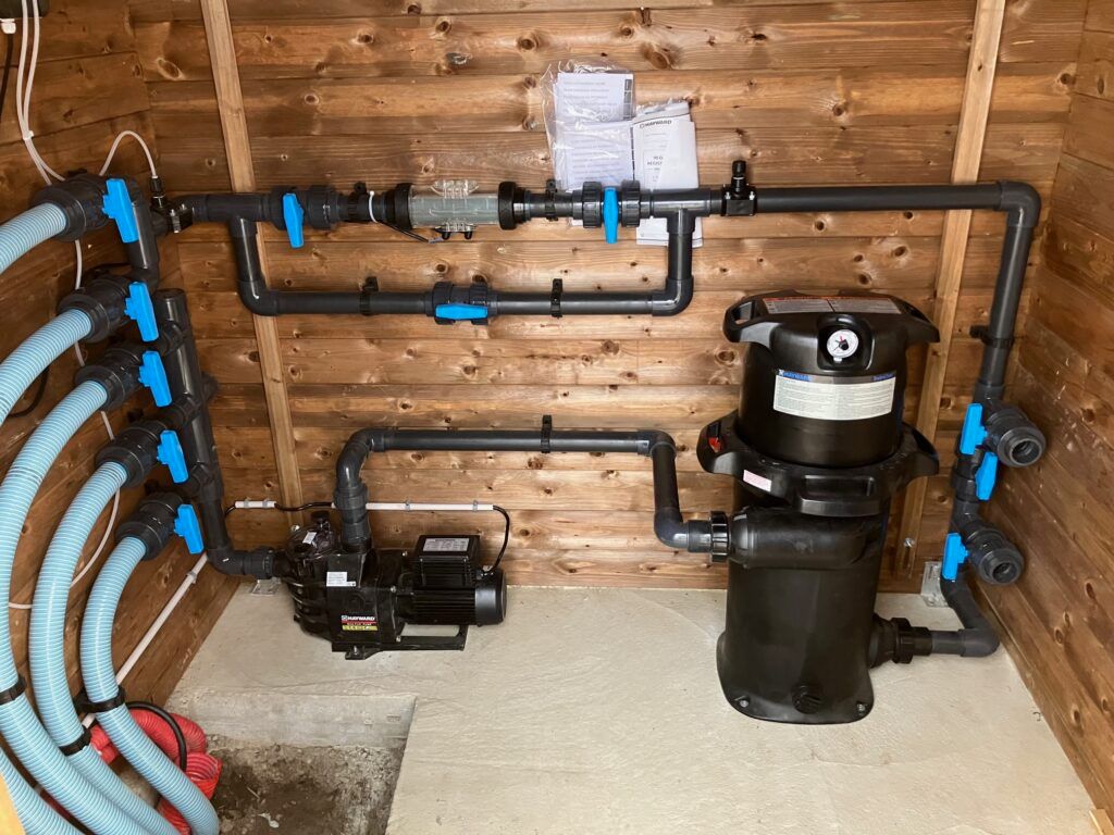 Pompe et système de filtration de piscine installés à l'intérieur d'une structure en bois avec des tuyaux et des vannes de plomberie bleus.