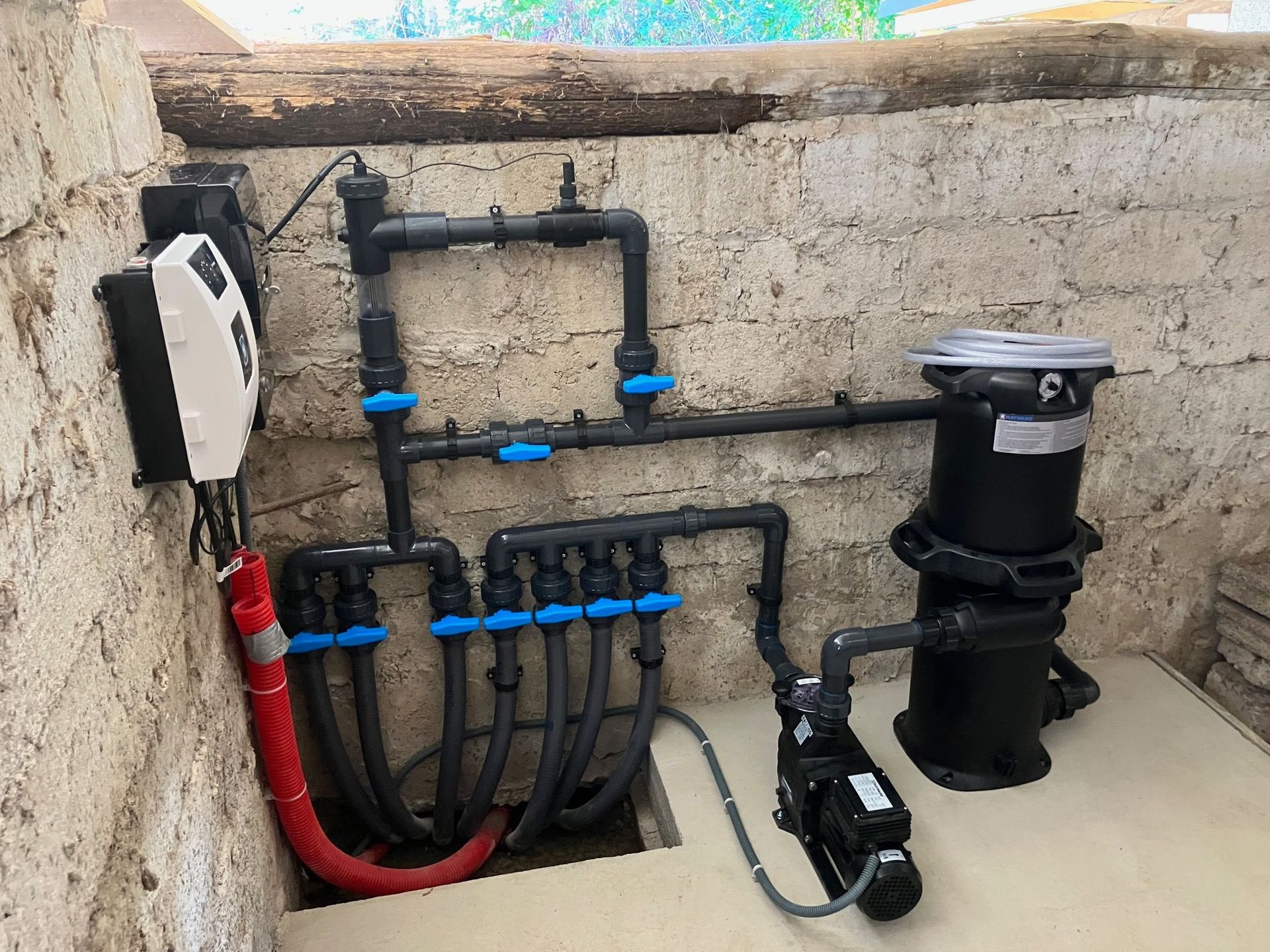Équipement de pompe de piscine installé dans un enclos en béton avec des tuyaux noirs, des vannes bleues, un filtre et un moteur de pompe.