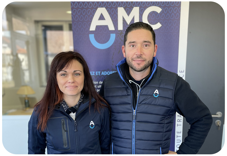 Deux personnes se tiennent devant une banderole bleue portant la marque « AMC », toutes deux vêtues de vestes sombres ornées du logo AMC.