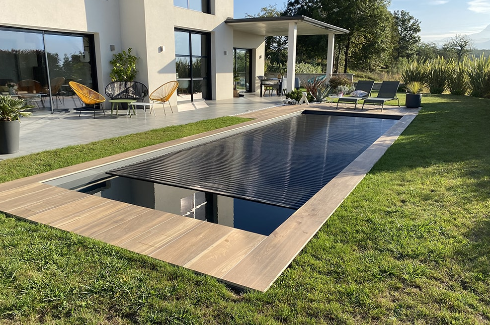 Une piscine rectangulaire avec une couverture sombre et fermée, entourée d'une terrasse et d'une pelouse, jouxtant une maison moderne.