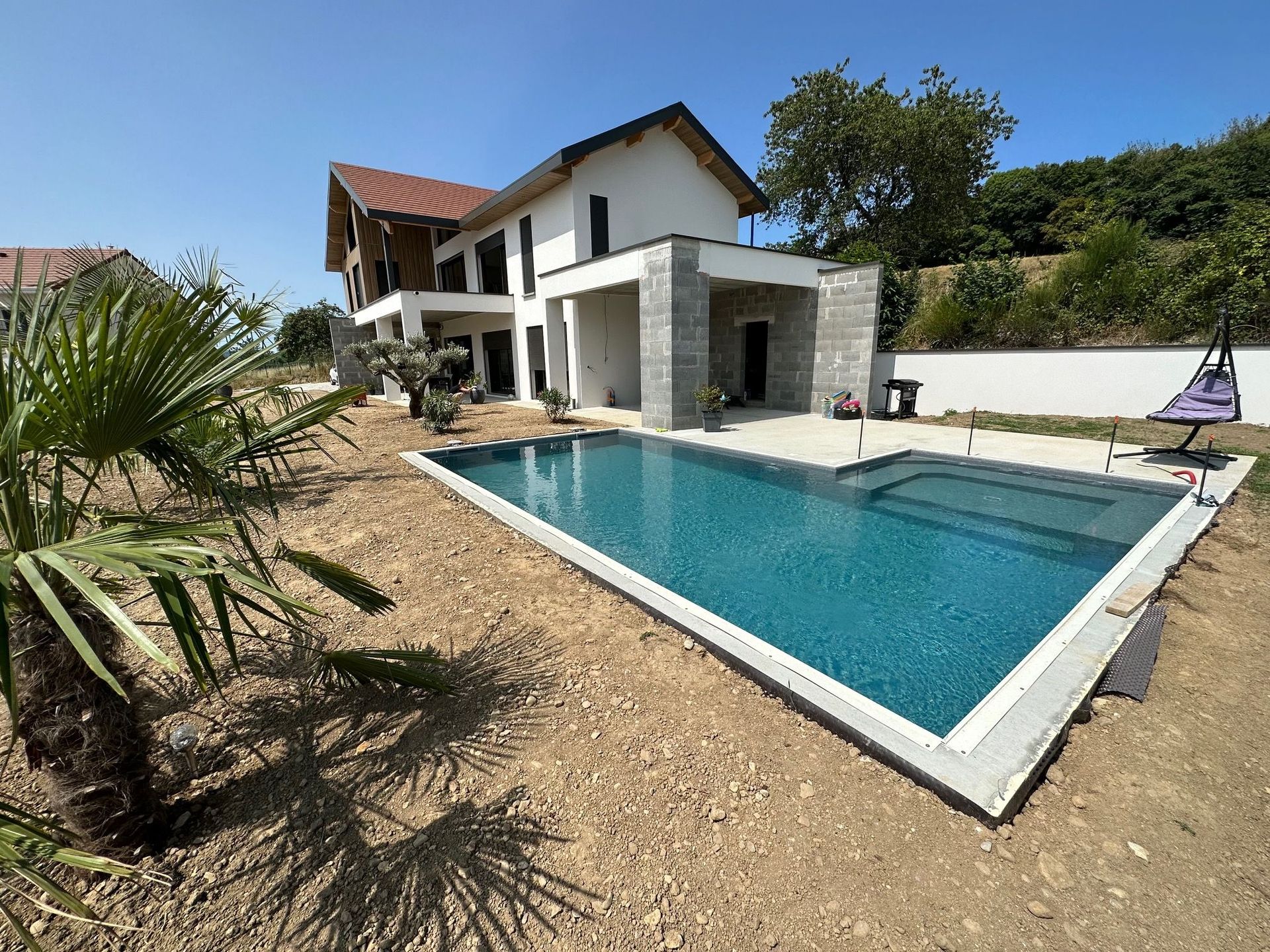 Maison blanche moderne avec des éléments en pierre et une piscine rectangulaire située dans une cour gravillonnée sous un ciel d'un bleu éclatant.