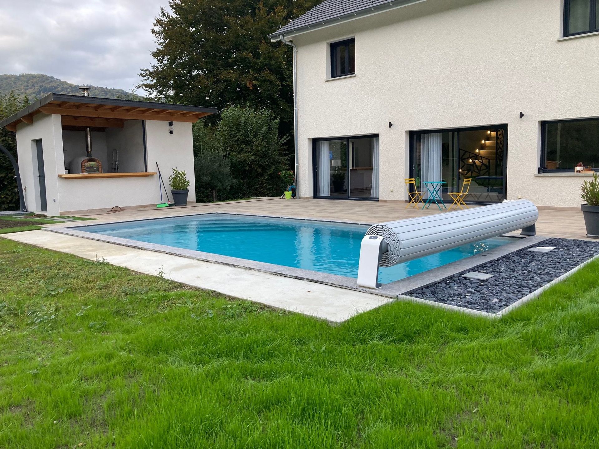 Une maison moderne et un petit abri extérieur à côté d'une piscine avec une bâche de sécurité partiellement enroulée dans une cour.