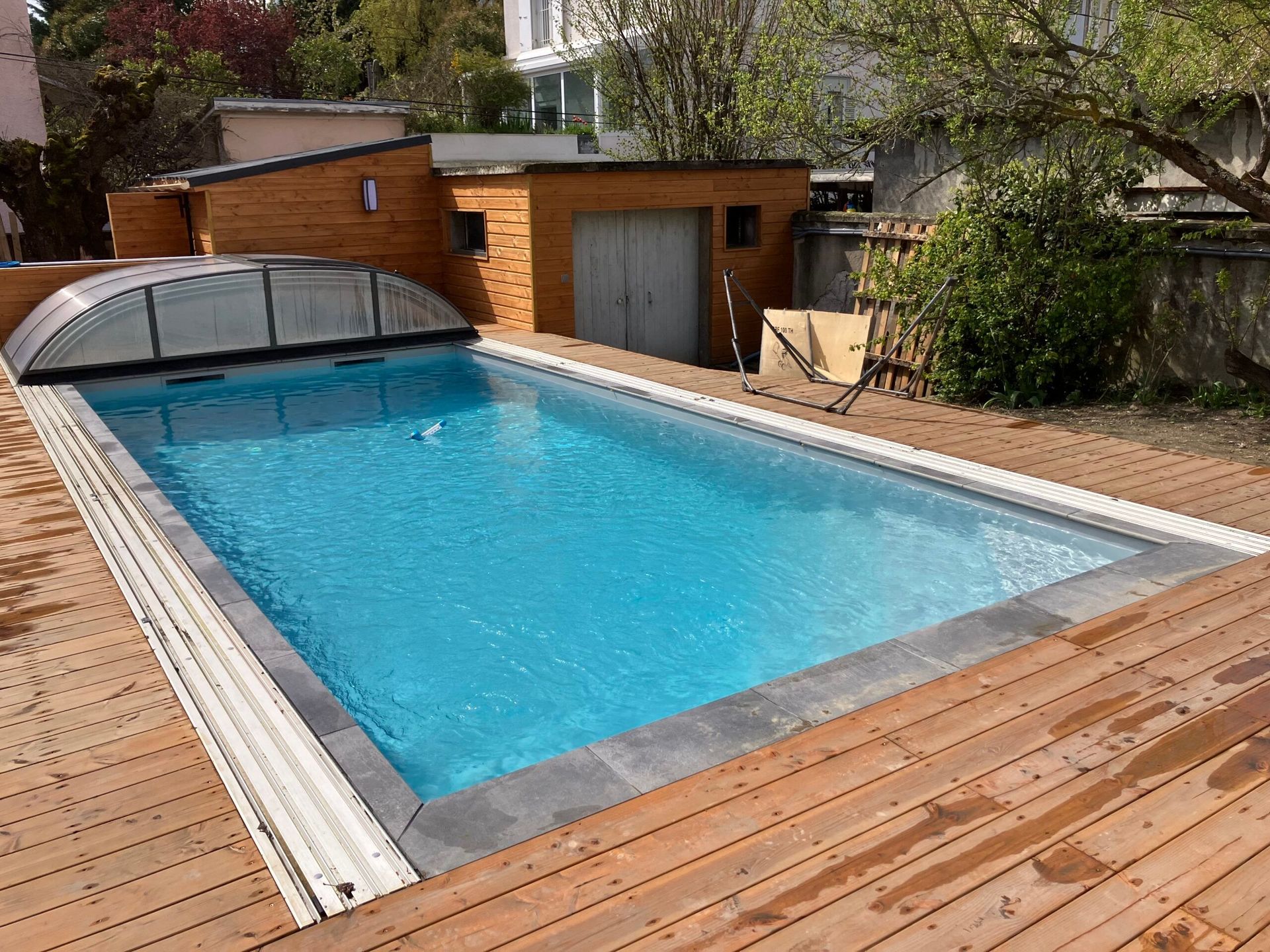 Une piscine rectangulaire dans un jardin, avec une couverture partiellement fermée, entourée d'une terrasse en bois et d'un petit abri de jardin.
