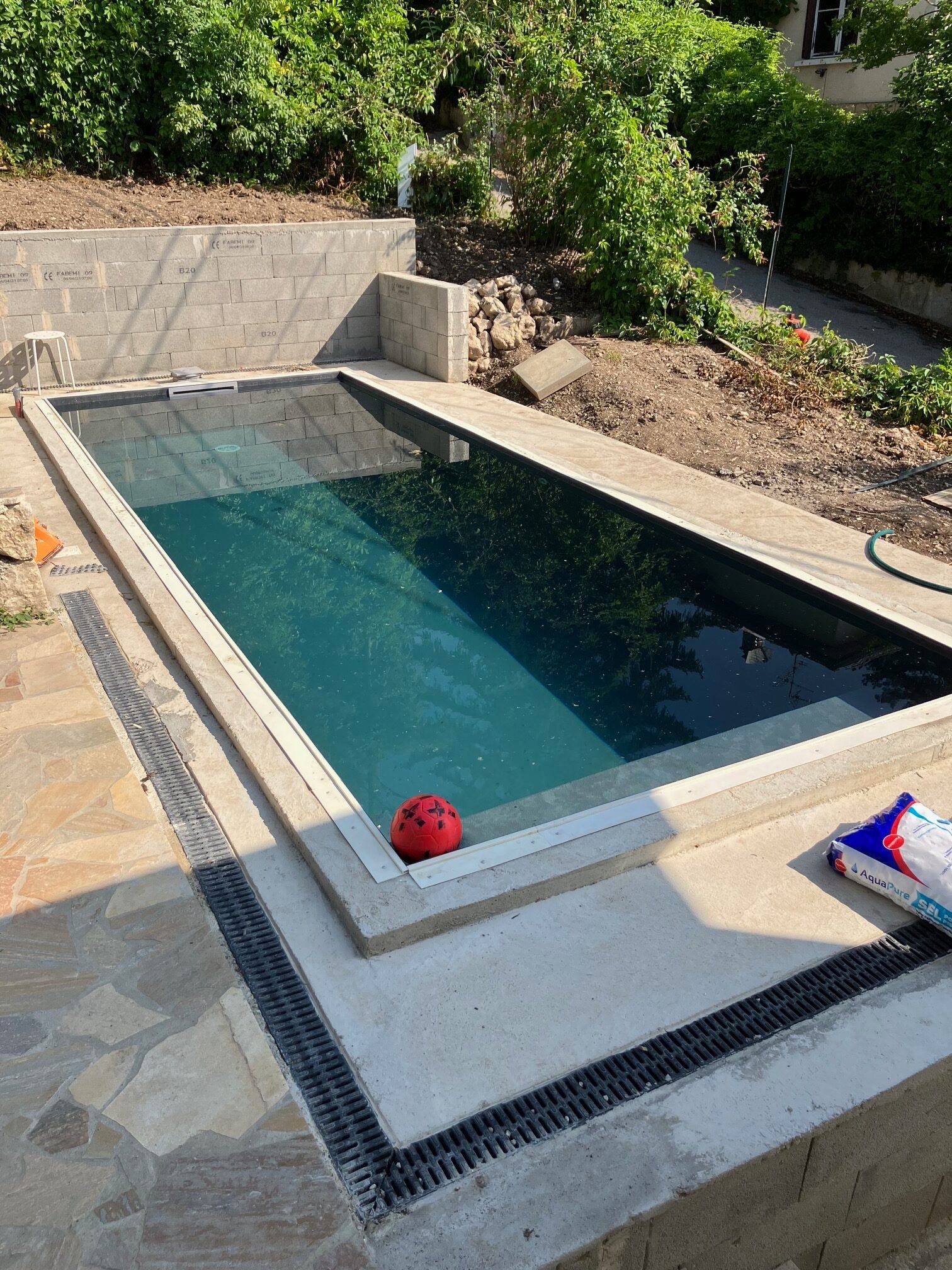 Une piscine extérieure rectangulaire aux eaux sombres et à la terrasse en béton en construction, entourée de verdure.
