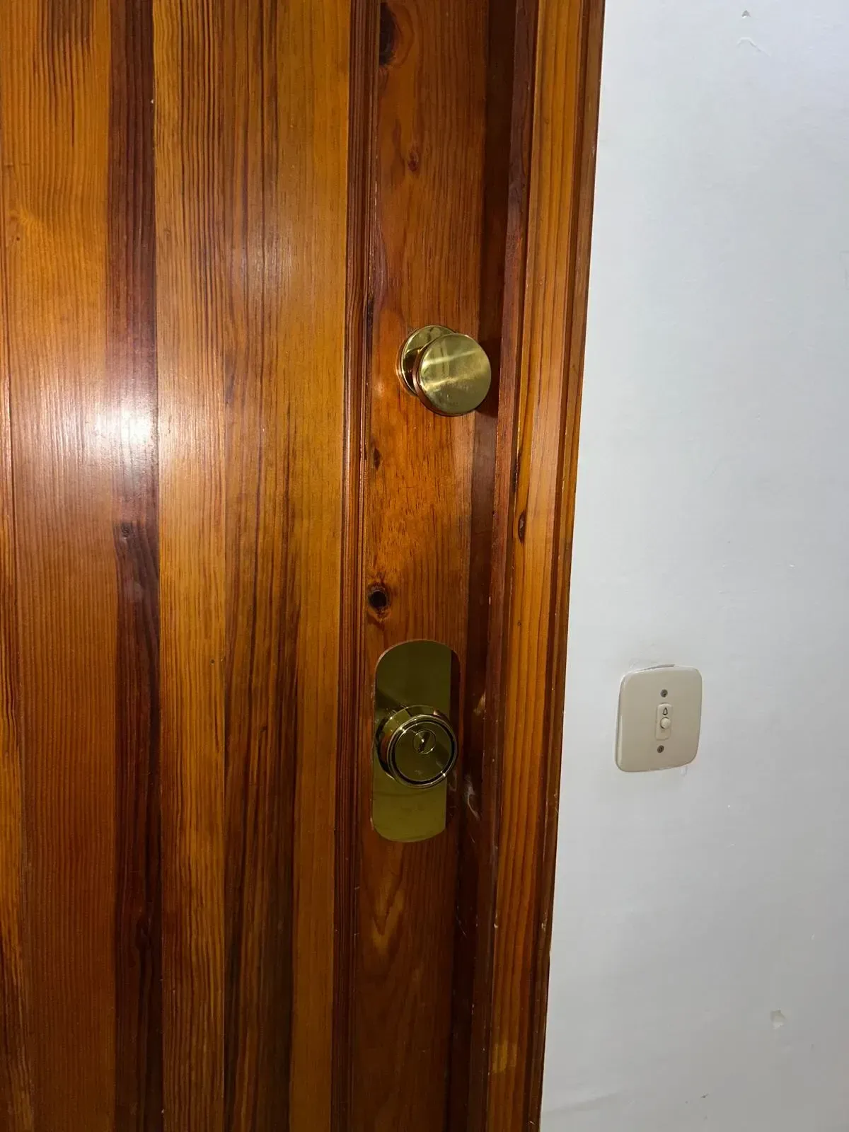 Puerta de madera con cerrojo y pomo de latón junto a una pared blanca y un interruptor de luz.