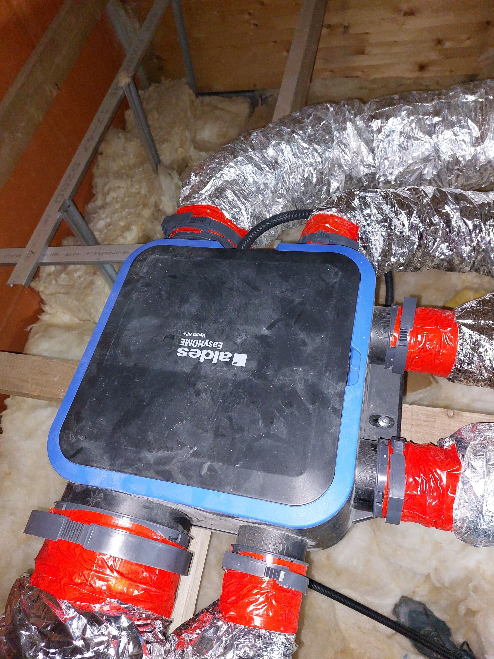 Unité de ventilation rectangulaire noire et bleue dans les combles, reliée à un réseau de conduits recouverts de tissu rouge et argenté.