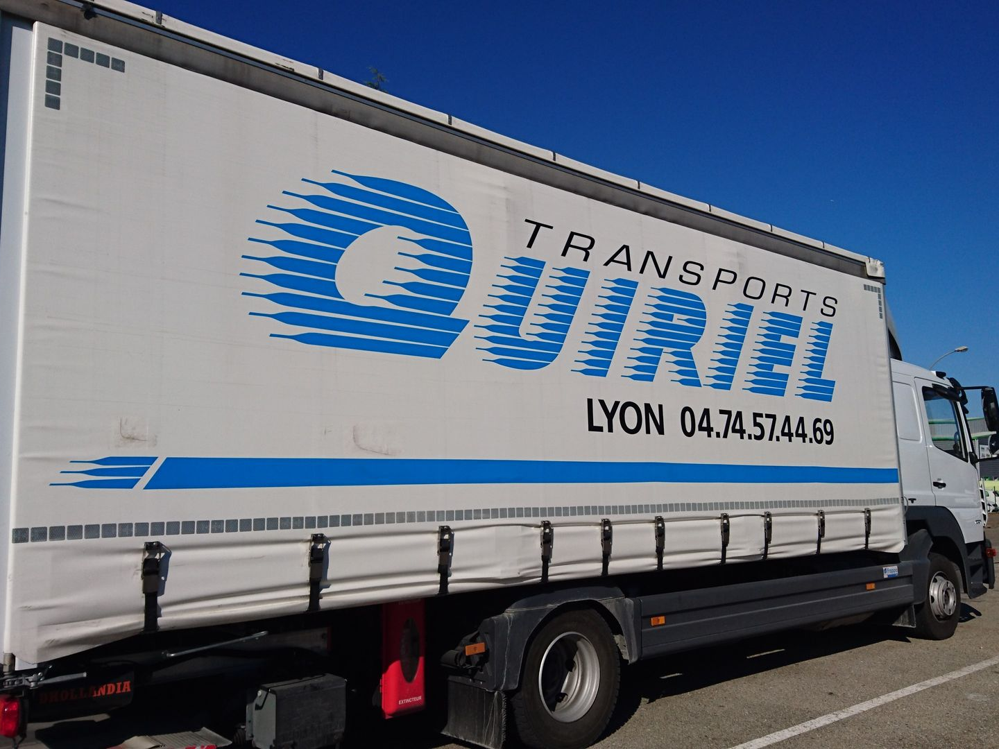 Camion blanc avec TRANSPORTS QUIRIEL LYON et numéro de téléphone sur le côté, logo bleu, sous un ciel clair.
