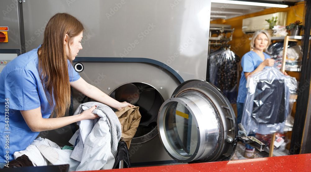 Eine Frau belädt in einem Waschsalon eine Frontlader-Waschmaschine mit Wäsche; im Hintergrund ist eine weitere Person zu sehen.