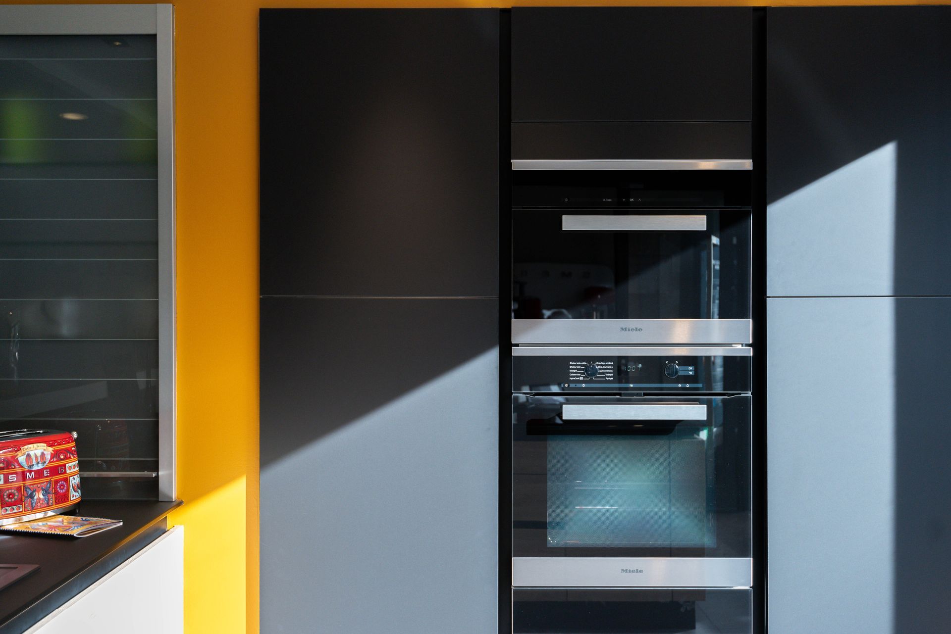 Cuisine moderne avec fours encastrés noirs et placards gris, le tout adossé à un mur jaune vif.