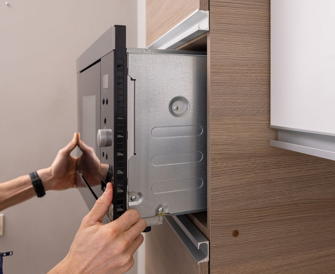 Des mains glissent un four à micro-ondes encastrable dans un meuble en bois d'une cuisine.