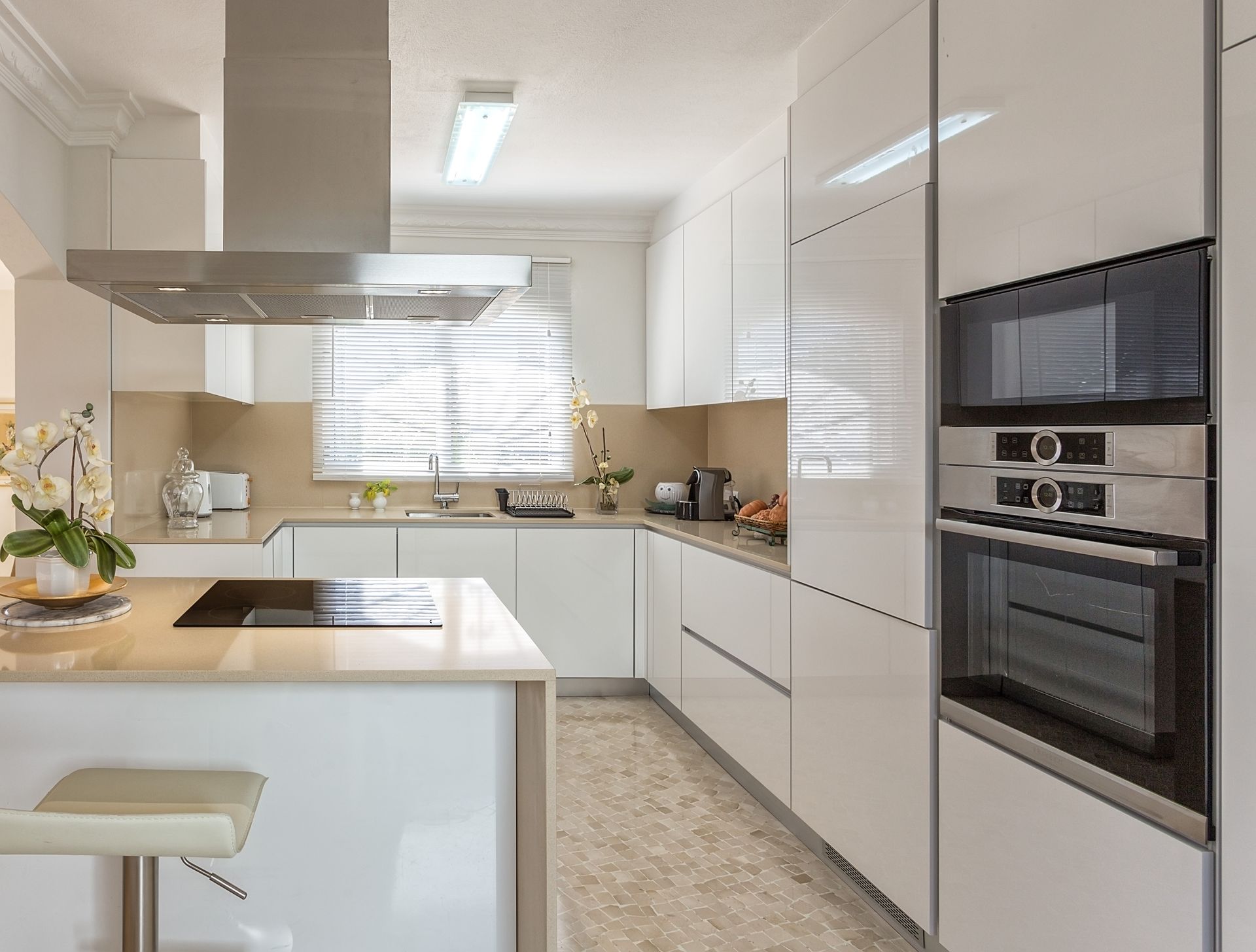 Cuisine moderne blanche avec îlot central, four encastré, hotte en inox et sol carrelé.