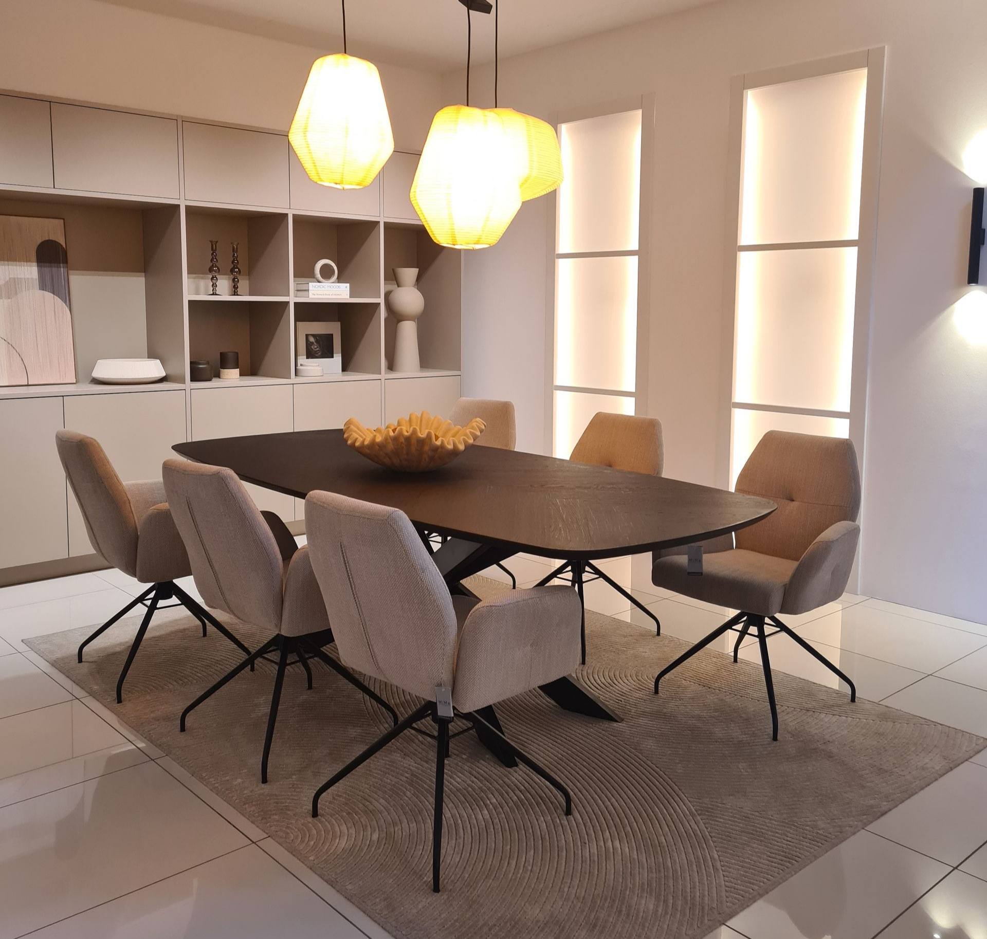 Salle à manger moderne avec une table en bois, six chaises, des suspensions lumineuses et des étagères intégrées.