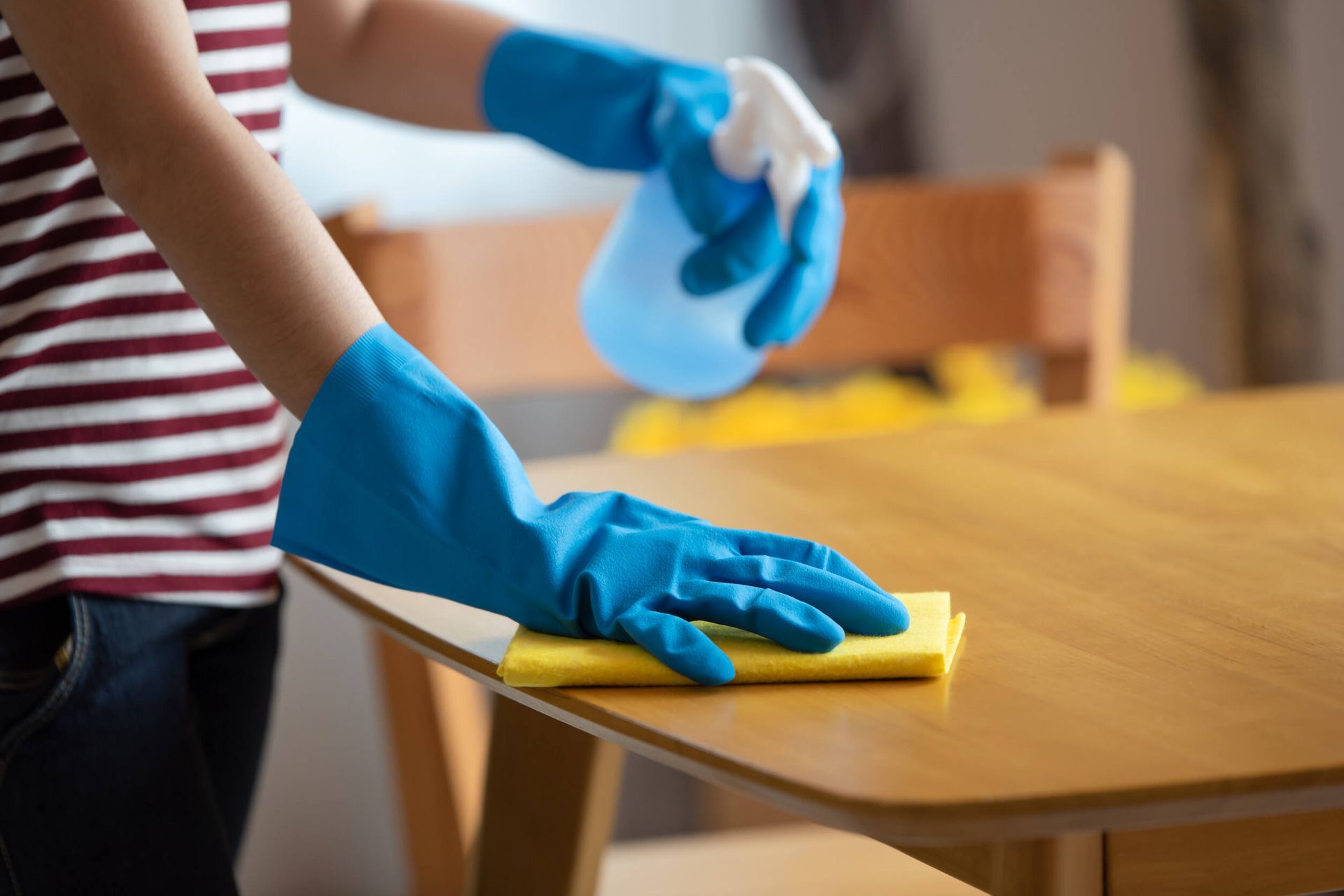 Una persona que lleva guantes azules está limpiando una mesa de madera con un paño y una botella de spray.