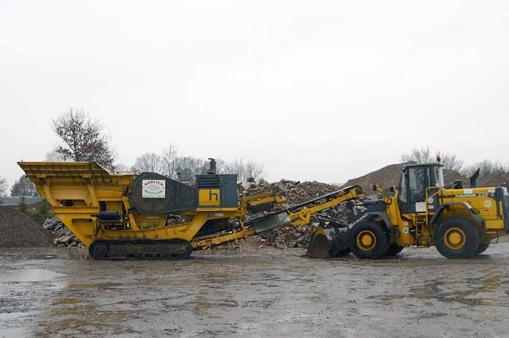Gelbe Schwermaschinen verarbeiten Schutt an einem nassen, grauen Tag, unterstützt von einem Radlader.