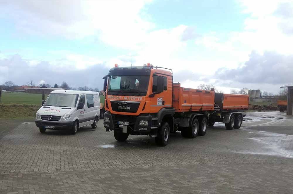 Ein orangefarbener Muldenkipper und ein weißer Lieferwagen parken auf einem gepflasterten Parkplatz unter bewölktem Himmel.