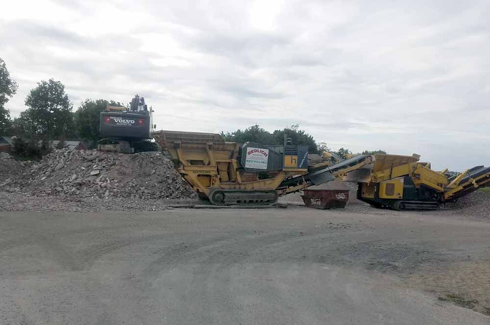 Baustelle mit Baumaschinen, darunter ein gelber Brecher und ein Bagger, unter bewölktem Himmel.